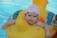 A baby with a pink floral-patterned hat is sitting in a yellow inflatable pool toy in the shape of a duck. The water in the pool is a clear blue, and the baby is smiling.