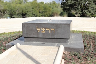 A dark gray rectangular monument with gold-colored Hebrew inscriptions on its front side stands in an outdoor setting. It is surrounded by a neatly maintained garden area with green shrubs and trees in the background. The pathway leading to the monument is paved with light-colored stones.