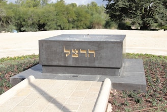 A dark gray rectangular monument with gold-colored Hebrew inscriptions on its front side stands in an outdoor setting. It is surrounded by a neatly maintained garden area with green shrubs and trees in the background. The pathway leading to the monument is paved with light-colored stones.