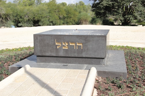 A dark gray rectangular monument with gold-colored Hebrew inscriptions on its front side stands in an outdoor setting. It is surrounded by a neatly maintained garden area with green shrubs and trees in the background. The pathway leading to the monument is paved with light-colored stones.