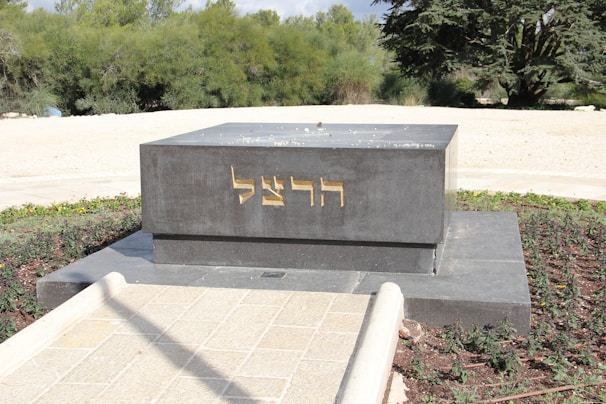 A dark gray rectangular monument with gold-colored Hebrew inscriptions on its front side stands in an outdoor setting. It is surrounded by a neatly maintained garden area with green shrubs and trees in the background. The pathway leading to the monument is paved with light-colored stones.