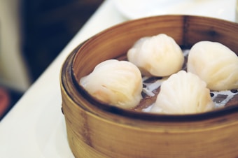 A bamboo steamer containing four translucent dumplings, often known as har gow, on a white surface. The dumplings have a slightly shiny appearance and are arranged neatly in the steamer basket.