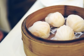 A bamboo steamer containing four translucent dumplings, often known as har gow, on a white surface. The dumplings have a slightly shiny appearance and are arranged neatly in the steamer basket.