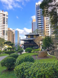 A traditional Japanese-style building surrounded by lush green gardens and neatly trimmed shrubs. Tall modern buildings tower in the background under a bright blue sky with scattered clouds. A small pond is visible to the right, adding to the calm ambiance of the setting.