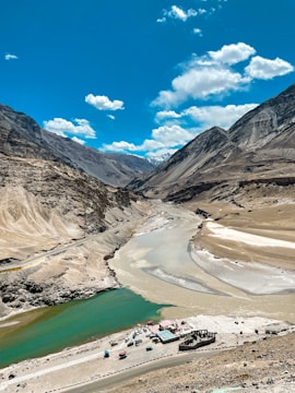 A picturesque landscape featuring a river flowing through a mountainous region. The river has a unique confluence where turquoise and brown waters meet, surrounded by rugged, arid mountains under a clear, bright blue sky with scattered clouds. At the riverbank, there are small buildings and parked vehicles, indicating a human presence amidst the vast natural scenery.