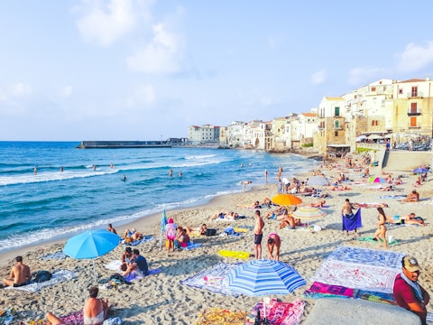 A lively beach scene with numerous people relaxing on the sand, sunbathing, and enjoying the sea. Brightly colored umbrellas and towels are scattered across the sandy shore. In the background, charming old buildings with balconies overlook the beach. The sea is calm, and a few people are swimming or standing in the shallow water.