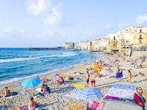 A lively beach scene with numerous people relaxing on the sand, sunbathing, and enjoying the sea. Brightly colored umbrellas and towels are scattered across the sandy shore. In the background, charming old buildings with balconies overlook the beach. The sea is calm, and a few people are swimming or standing in the shallow water.