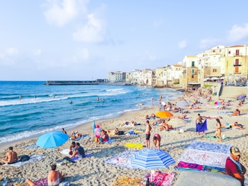 A lively beach scene with numerous people relaxing on the sand, sunbathing, and enjoying the sea. Brightly colored umbrellas and towels are scattered across the sandy shore. In the background, charming old buildings with balconies overlook the beach. The sea is calm, and a few people are swimming or standing in the shallow water.