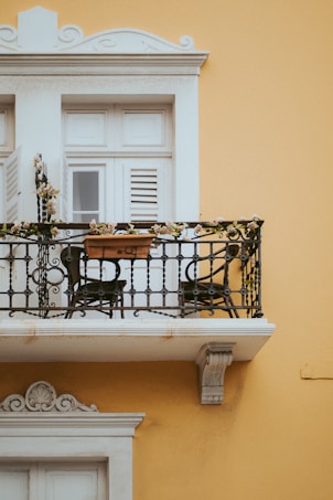 A charming balcony with intricate wrought iron railing adorned with flowers. The backdrop features a vibrant yellow wall and a white-framed shuttered window. A small potted plant adds a touch of nature to the setting.
