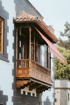 A picturesque wooden balcony with intricate carvings, supported by detailed columns, extends from a building with a checkered pattern of black and white tiles. A red retractable awning provides shade, and greenery is visible in the background.