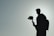 A silhouette of a person holding a camera on a gimbal. The background is a gradient of light gray to blue, creating a muted and minimalist atmosphere. The person wears a backpack, adding to the sense of travel or adventure.