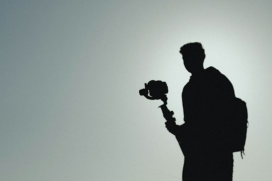 A silhouette of a person holding a camera on a gimbal. The background is a gradient of light gray to blue, creating a muted and minimalist atmosphere. The person wears a backpack, adding to the sense of travel or adventure.