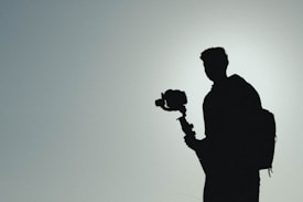 A silhouette of a person holding a camera on a gimbal. The background is a gradient of light gray to blue, creating a muted and minimalist atmosphere. The person wears a backpack, adding to the sense of travel or adventure.