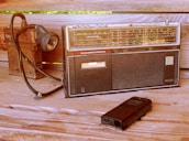 A vintage radio recorder with a cassette player and AM/FM tuning dials is positioned on a wooden surface. Beside it is an old-fashioned flashlight with a coiled cable and a portable cassette player, adding to the retro ambiance.