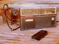 A vintage radio recorder with a cassette player and AM/FM tuning dials is positioned on a wooden surface. Beside it is an old-fashioned flashlight with a coiled cable and a portable cassette player, adding to the retro ambiance.