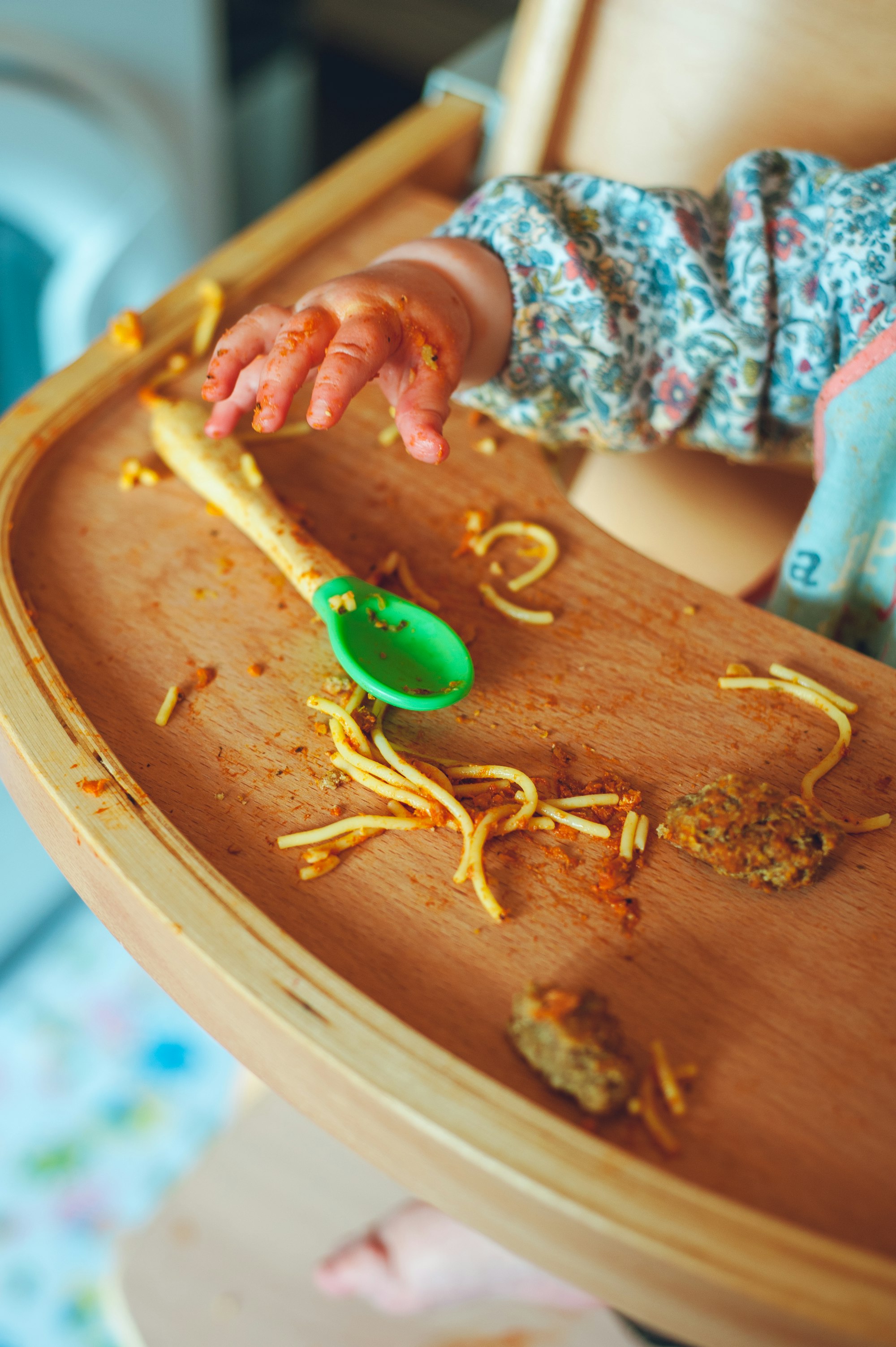 Toddler in a wooden high chair eating spaghetti in a tomato sauce 