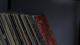 A collection of vinyl records neatly lined up on a shelf, showcasing various album covers with different colors and designs. The lighting is dim, casting a muted and nostalgic atmosphere.