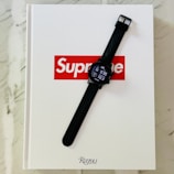 A black digital watch with a silicone strap is positioned on top of a white book with the red and white Supreme logo on the cover. The book is set on a marble surface and its publisher is Rizzoli New York.