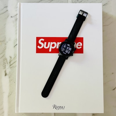 A black digital watch with a silicone strap is positioned on top of a white book with the red and white Supreme logo on the cover. The book is set on a marble surface and its publisher is Rizzoli New York.