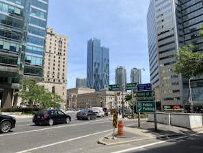 A busy urban intersection surrounded by modern high-rise buildings with glass facades. Several road signs are visible, directing traffic towards York St South, Front St Eastbound, and Front St Westbound. Vehicles including cars and vans move along the streets. The backdrop features a prominent blue skyscraper, and trees add greenery to the scene.