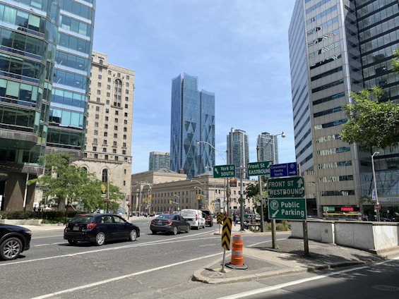 A busy urban intersection surrounded by modern high-rise buildings with glass facades. Several road signs are visible, directing traffic towards York St South, Front St Eastbound, and Front St Westbound. Vehicles including cars and vans move along the streets. The backdrop features a prominent blue skyscraper, and trees add greenery to the scene.