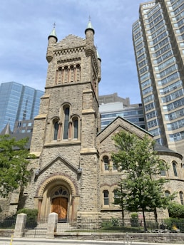 A historic stone church with ornate architectural features, including arched windows and a tall bell tower. The structure stands in contrast to the modern high-rise buildings surrounding it. Trees and a gated entrance are visible in the foreground.