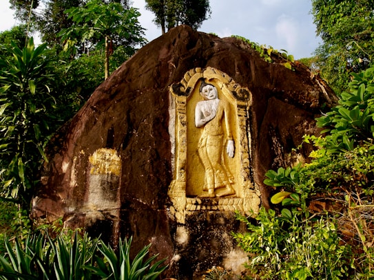 A carved statue of a figure embedded in a large rock, surrounded by lush green plants and trees. The statue is adorned with gold paint, standing in a graceful pose within an ornate frame carved into the rock.