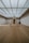 A minimalist interior room with wooden flooring and a large, geometric triangular structure as the centerpiece. The ceiling is made of glass panels that diffuse soft light throughout the space, creating a serene ambiance. The walls are plain and complement the simplicity of the design.