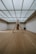 A minimalist interior room with wooden flooring and a large, geometric triangular structure as the centerpiece. The ceiling is made of glass panels that diffuse soft light throughout the space, creating a serene ambiance. The walls are plain and complement the simplicity of the design.