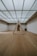 A minimalist interior room with wooden flooring and a large, geometric triangular structure as the centerpiece. The ceiling is made of glass panels that diffuse soft light throughout the space, creating a serene ambiance. The walls are plain and complement the simplicity of the design.