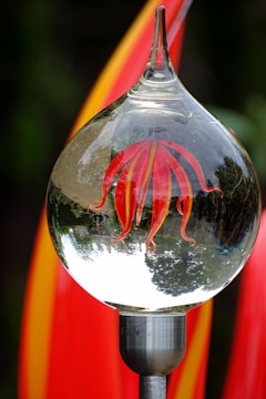 A clear glass orb with red and yellow abstract shapes inside is mounted on a metallic stand. The orb reflects an inverted image of a garden with trees, creating a distorted view. Bright red and orange streaks in the background complement the colors inside the orb.