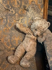 A well-worn teddy bear is slumped in an armchair with an intricate floral pattern. The bear's fur is frayed and faded, giving it a vintage appearance. The chair's fabric features ornate designs, predominantly in earthy tones.