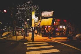A nighttime urban scene features a corner restaurant named 'Middle East' with a warmly lit yellow awning and neon signs. Several people can be seen walking or standing nearby, and the street is adorned with string lights that illuminate the area. Sidewalk seating is partially visible with a few patrons.