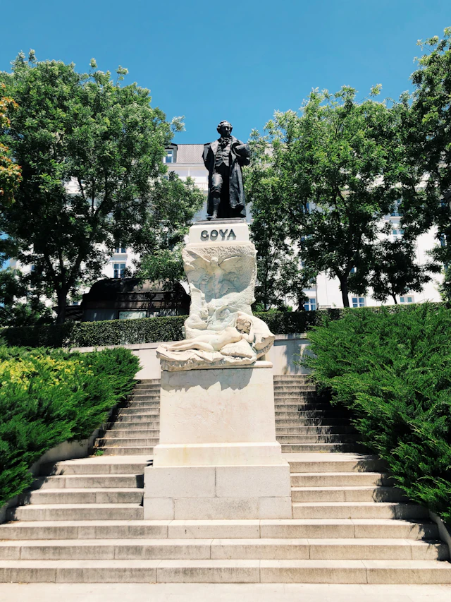 The bronze Goya monument on its stone pedestal in the gardens outside the Prado museum, flanked by trimmed hedges and a flight of steps.