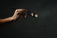 A hand delicately holding a piece of sushi with chopsticks against a dark, moody background. The lighting highlights the texture of the rice and the topping.