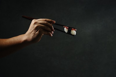 A hand delicately holding a piece of sushi with chopsticks against a dark, moody background. The lighting highlights the texture of the rice and the topping.