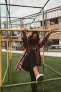A person wearing a red plaid shirt and black pants is interacting with a jungle gym structure in an outdoor playground. The individual’s face is partially covered with a mask and they are holding onto the yellow bars, while lifting one leg as if climbing or balancing. The background shows a grassy field and part of a building with windows and railing.
