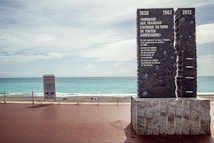 A memorial plaque stands prominently on a promenade overlooking a blue ocean. The plaque is made of dark stone with carved text commemorating the contribution of North African French citizens. The sky is cloudy, adding a serene yet somber atmosphere.