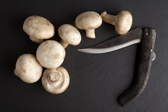 A group of fresh white mushrooms is arranged on a dark slate surface next to a sleek, curved knife with a wooden handle. The composition of the items suggests a cooking or culinary context.