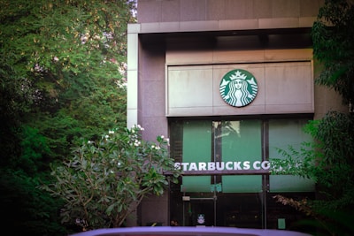 The entrance of a building features prominent branding with the recognizable Starbucks logo above the doors. Surrounding the structure are lush, green trees and plants, adding a natural ambiance.