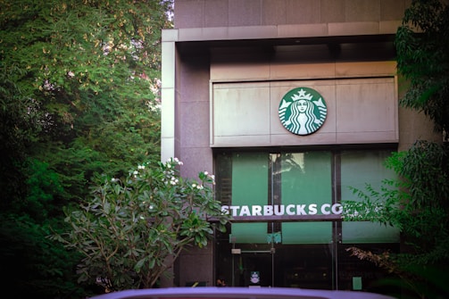 The entrance of a building features prominent branding with the recognizable Starbucks logo above the doors. Surrounding the structure are lush, green trees and plants, adding a natural ambiance.