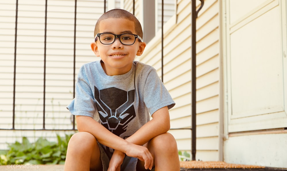 boy in gray crew neck t-shirt wearing black framed eyeglasses sitting on brown ground during