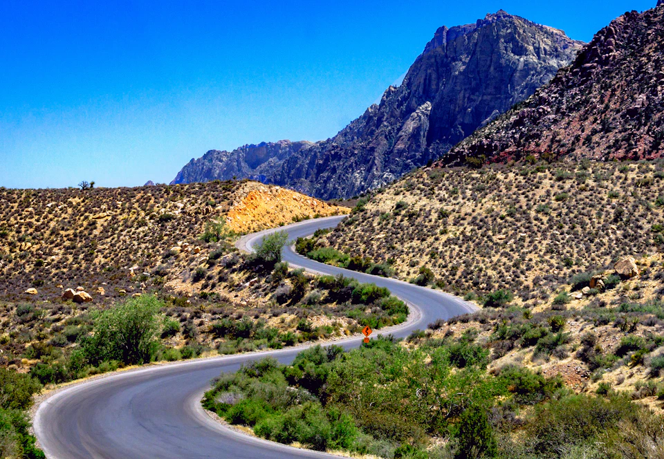 Winding desert road cuts through mountain range with sparse vegetation and rocky outcroppings
