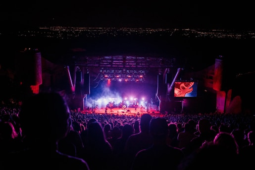 A large crowd attending a nighttime outdoor concert. The stage is illuminated with bright lights, featuring multiple musicians performing. A large screen to the right of the stage displays a close-up of a performer playing a guitar. The background consists of a dark sky, suggesting the concert is set in an open-air venue, possibly with a cityscape visible faintly in the distance.