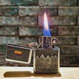 A metallic lighter with an embossed design of a cityscape is lit, displaying a vibrant blue and orange flame. The lid is open, revealing the words 'Germany' and 'Deutschland' alongside a German flag. The background consists of a textured stone wall, and the lighter is resting on a wooden surface.