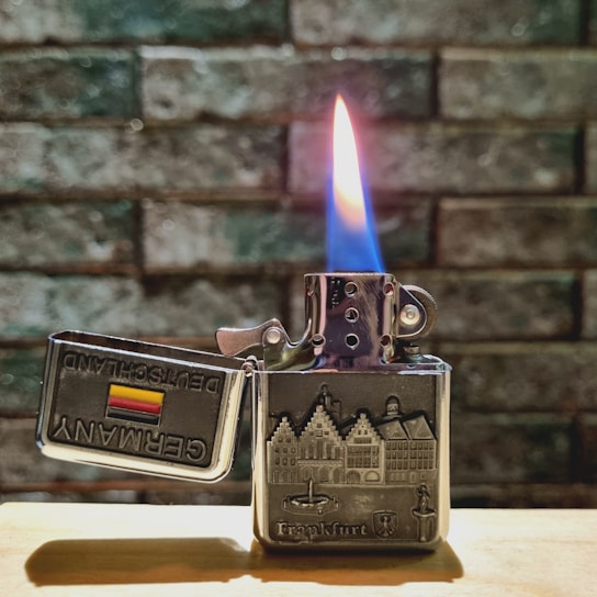 A metallic lighter with an embossed design of a cityscape is lit, displaying a vibrant blue and orange flame. The lid is open, revealing the words 'Germany' and 'Deutschland' alongside a German flag. The background consists of a textured stone wall, and the lighter is resting on a wooden surface.