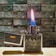A metallic lighter with an embossed design of a cityscape is lit, displaying a vibrant blue and orange flame. The lid is open, revealing the words 'Germany' and 'Deutschland' alongside a German flag. The background consists of a textured stone wall, and the lighter is resting on a wooden surface.