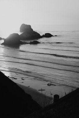A serene coastal scene with gentle waves rolling onto a sandy beach. Rock formations jut out from the sea, and the silhouette of a person holding a surfboard is visible near the water&rsquo;s edge. The landscape is captured in monochrome, creating a tranquil and timeless atmosphere.