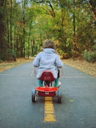 A child wearing a fluffy jacket is riding a tricycle down a paved path, surrounded by lush green trees. The path is marked with a yellow dashed line, and fallen leaves are scattered along the sides.