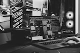 A monochrome image featuring a workspace with a MacBook Pro laptop displaying lines of code on its screen. Nearby, there's a second monitor also showing more code. In the background, a desktop computer with illuminated fans is visible, along with a hand on the laptop keyboard.